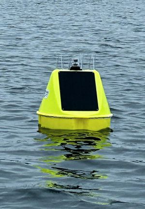 Yellow buoy with a dark solar panel and small antenna mast floating in rippling gray-blue water, reflecting a greenish tint below the surface.