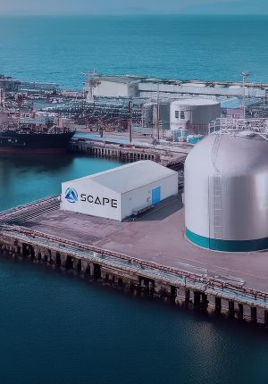 Aerial view of an industrial port facility with a white warehouse bearing the SCAPE logo next to a large domed storage tank, with a docked ship and ocean beyond.
