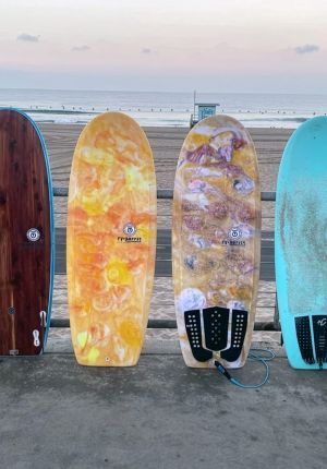 Four surfboards leaning against a beach railing at sunrise, two with swirled orange and marbled resin finishes made from recycled material.