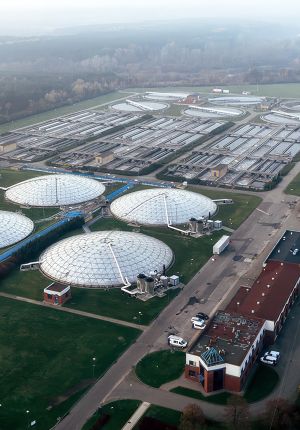 Aerial view of a large wastewater treatment plant with three white domed digesters and rows of rectangular settling basins in misty morning light.