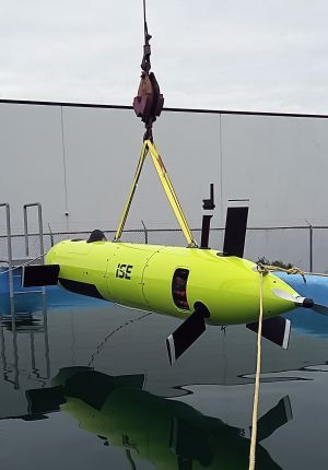 A bright yellow-green ISE autonomous underwater vehicle suspended from a crane above a test pool, with control fins and a propeller visible.