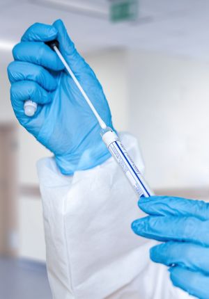 Gloved hands in blue nitrile gloves inserting a sterile swab into a labeled collection tube in a clinical setting.