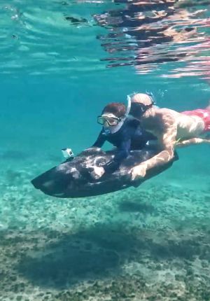 Two snorkelers underwater in clear turquoise water riding a black electric sea scooter fitted with an action camera above a sandy reef bottom.