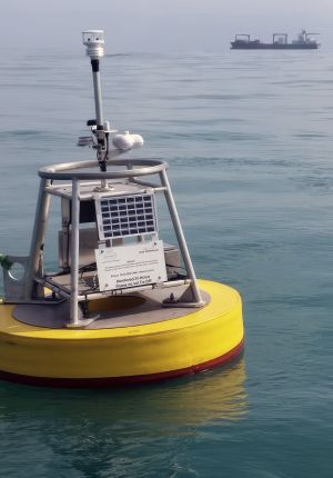 Yellow ocean monitoring buoy with solar panel and sensor mast, floating in open water with a cargo ship visible in the hazy background.