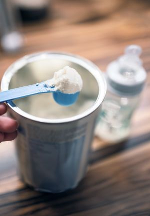 A blue plastic scoop of powdered infant formula held above an open canister, with a baby bottle blurred in the background on a wooden table.