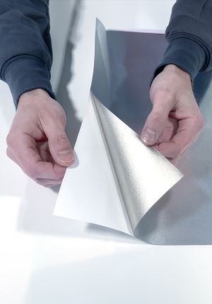 Two hands peeling back a corner of a large silver-grey thermal film sheet on a white surface, revealing a reflective metallic underside.