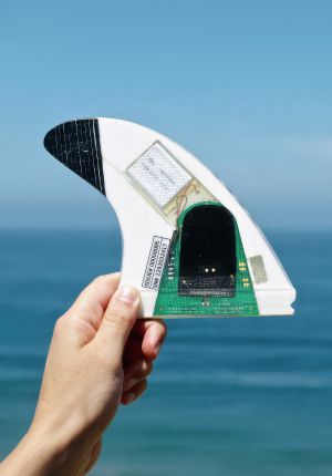 A hand holding up a white surfboard fin with embedded electronics — a visible circuit board, battery, and serial number label — against a blue ocean backdrop.