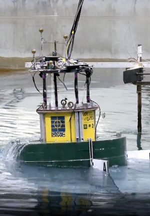 A scale model vessel with a yellow superstructure and instrumentation mast undergoing testing in a tow tank, pushing a bow wave.
