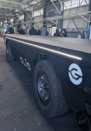 A large black GLID autonomous flatbed transport vehicle with off-road tires displayed indoors at a trade event, with attendees talking in the background.