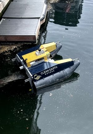 A navy and yellow Clean Earth Rovers catamaran drone operating in debris-strewn harbor water next to a floating dock.