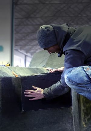 Worker in a beanie and hoodie smoothing dark composite material over the hull of a boat-shaped mold in a workshop.