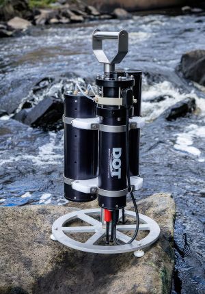 A black cylindrical DOT underwater sensor instrument mounted on a circular metal base plate, sitting on a rock beside rushing whitewater.