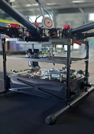 Close-up of an open drone frame on a workbench, showing exposed circuit boards, wiring, carbon fiber arms, and a GPS module.