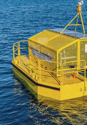 Close-up of a yellow floating platform equipped with solar panels and a mesh-enclosed equipment housing, anchored in open water with a warning sign for underwater lines.