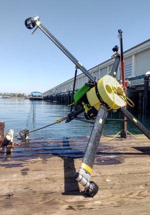An underwater robotic vehicle with carbon fiber arms, thrusters, and yellow buoyancy floats resting on a wooden pier beside calm harbor water.