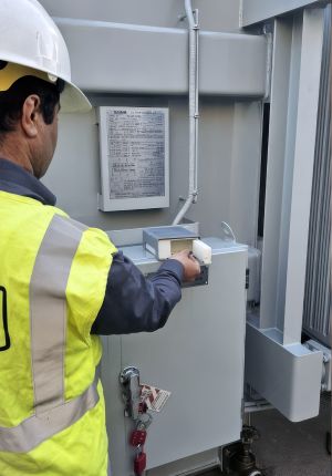 A worker in a white hard hat and high-visibility vest operating a control on a grey outdoor electrical cabinet with a lockout tag on the door handle.