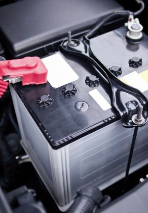 Close-up of a lead-acid vehicle battery installed in an engine bay, with a red terminal cover and metal hold-down bracket.