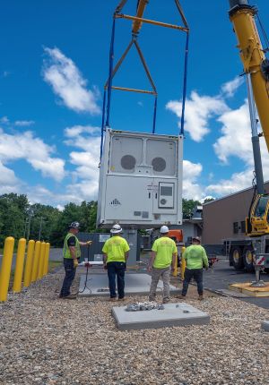 Four workers in high-visibility shirts and hard hats watching a crane lower a white containerized battery energy storage unit onto a concrete pad.