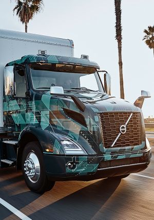 An electric box truck in teal geometric camouflage wrap driving on a palm-lined road.