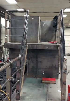 A large steel vessel hull section under construction inside a fabrication shop, with welded plating, stair railings, and tools on the deck.