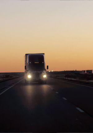 A semi truck with headlights on driving toward the camera on an open highway at dusk, silhouetted against an orange sunset sky.