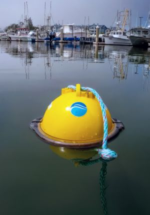 A yellow domed smart buoy with a blue wave logo floating in calm harbor water, tethered by braided rope, with fishing boats moored in the background.