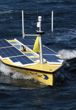 A yellow SeaTrac autonomous surface vessel covered in solar panels, with antennas and a sensor mast, cutting through dark blue open ocean.