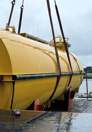 Large yellow cylindrical tank being lifted by crane straps at a dockside facility, likely a subsea or offshore energy component.